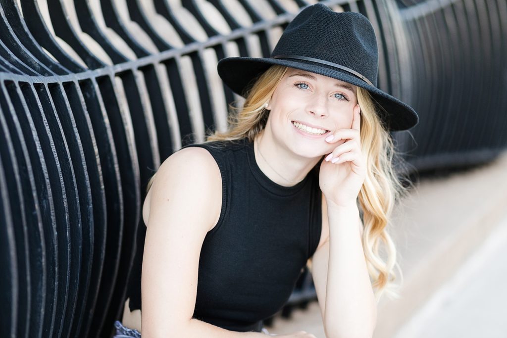 girl sitting against a black railing with a black shirt and black hat on smiling for her Eau Claire Senior Pictures