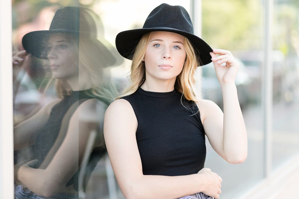 girl in a black shirt and black hat leaning against a glass wall for her Eau Claire Boho Senior Photos