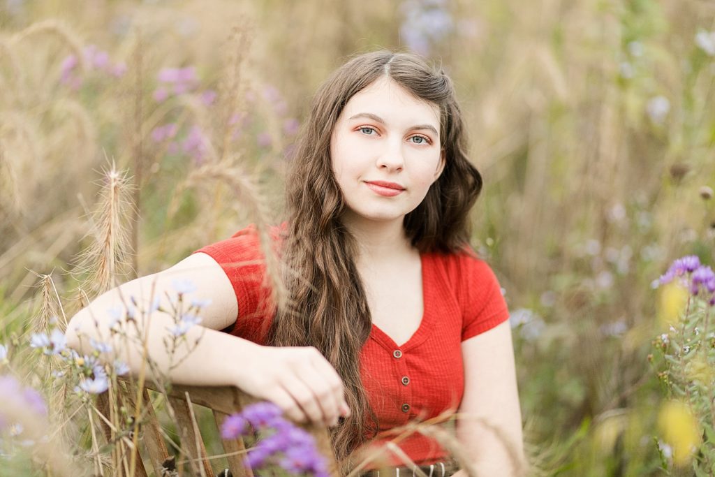 girl in an orange shirt in a field of fall flowers with purple and wheat for her Cadott High School senior photos
