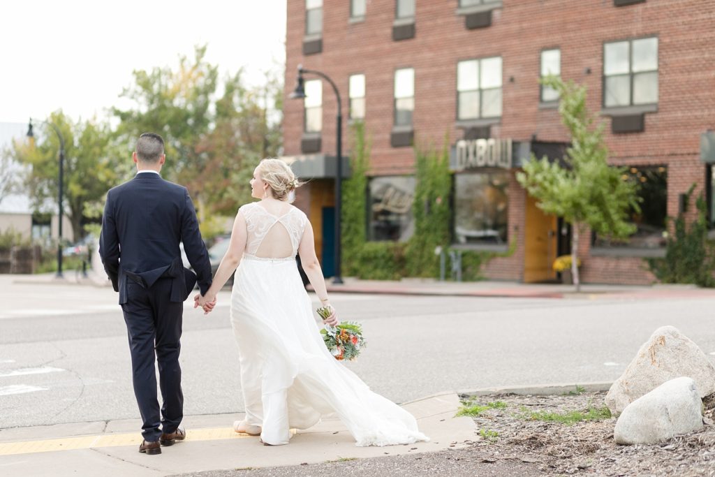 couple crossing the street with their wedding venue in the background at their wedding at the Oxbow Hotel in Eau Claire