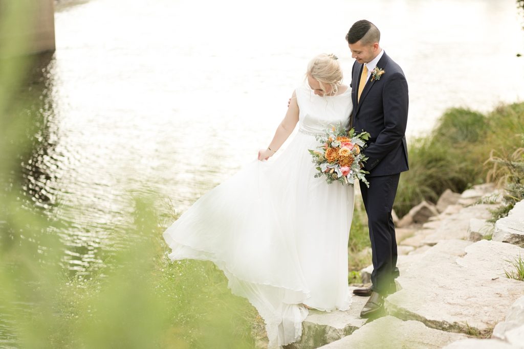 bride adjusting her dress near the river while the groom looks down at her at their wedding at the Oxbow Hotel in Eau Claire