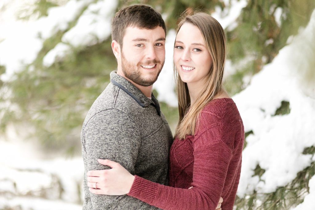 couple smiling at the camera tucked into some pine boughs for their winter Wisconsin engagement session