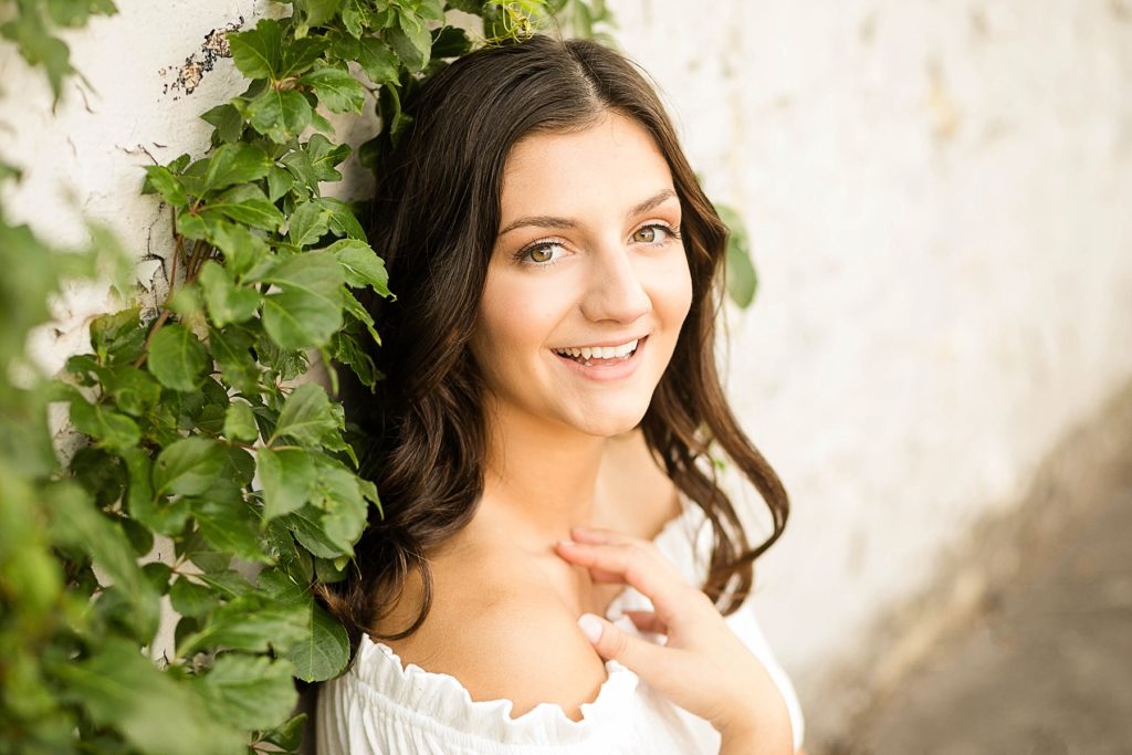girl sitting against a white wall with vines trailing down for her Bloomer summer senior pictures
