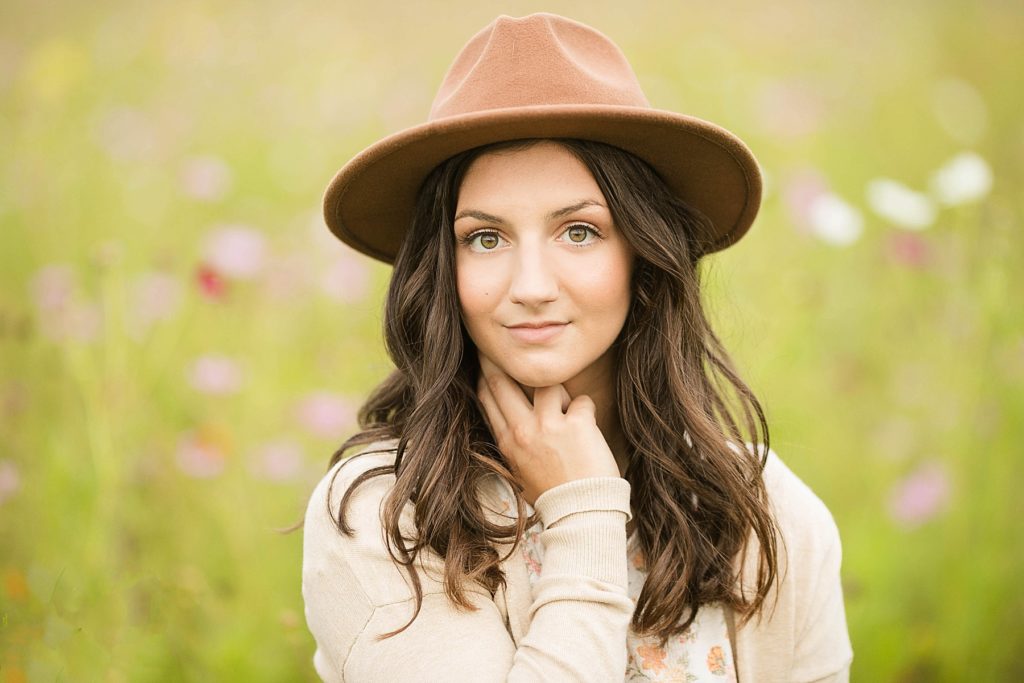 girl with a brown hat looking in to the camera sitting in a field of flowers for her senior photos in Chetek WI