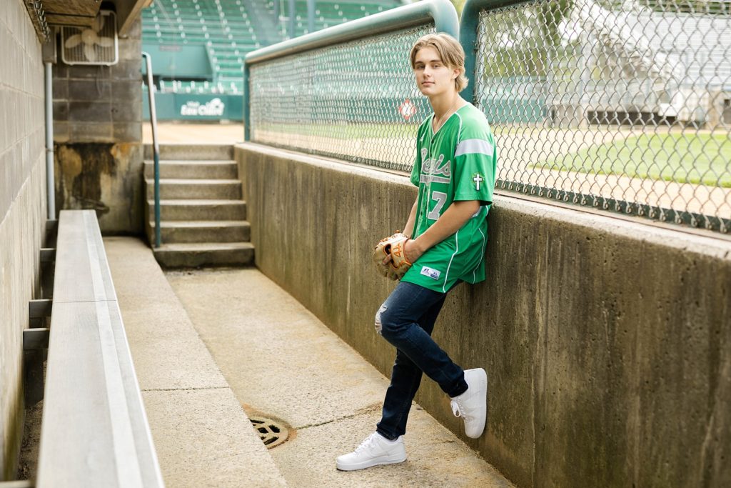 boy sitting in dugout with a green Regis High School baseball jersey on in Carson Park in Eau Claire for his senior photos