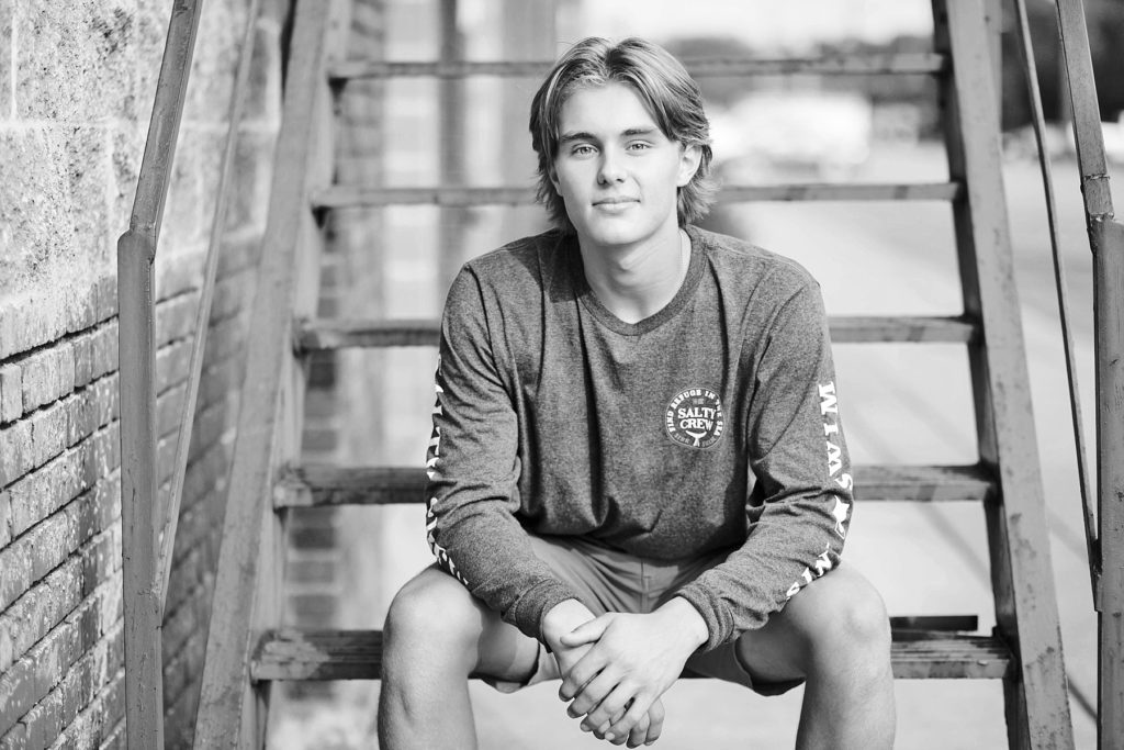 boy sitting on metal stairs in shorts in a black and white photo at Banbury Place in Eau Claire for his senior pictures