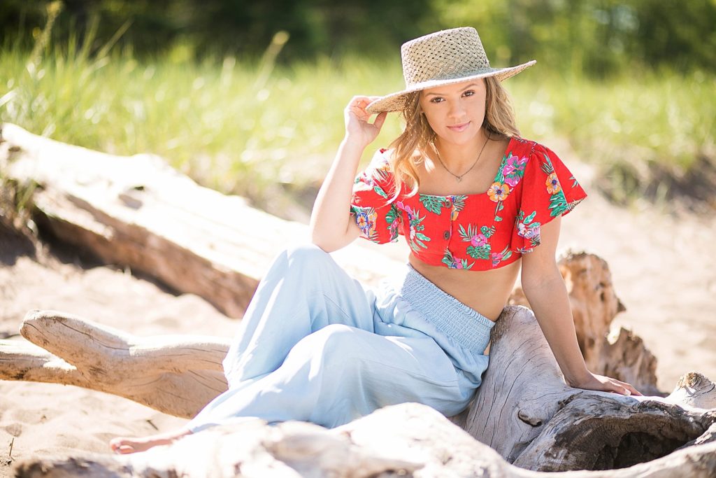 girl in a red floral crop top and denim pants with a Gigi Pip hat sits on Wisconsin Beach in Superior, WI for her senior photos