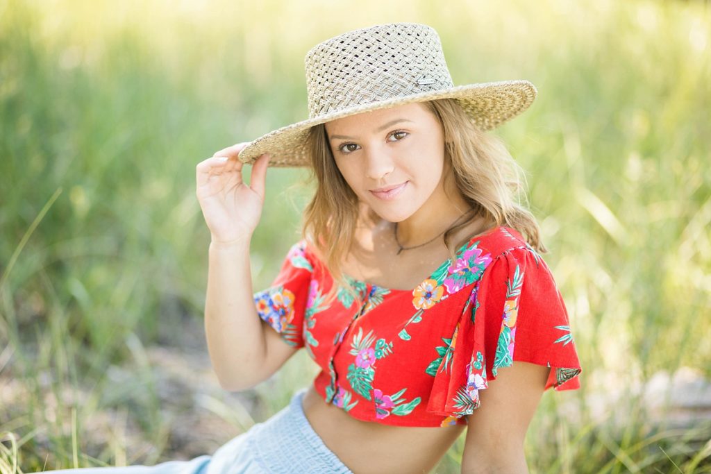 girl in a red floral crop top and denim pants with a Gigi Pip hat sits on Wisconsin Beach in Superior, WI for her senior photos