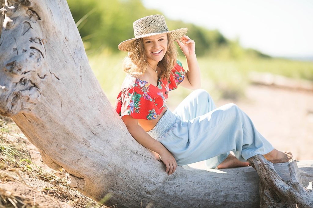 girl in a red floral crop top and denim pants with a Gigi Pip hat sits on a piece of driftwood at Wisconsin Beach in Superior, WI for her senior photos