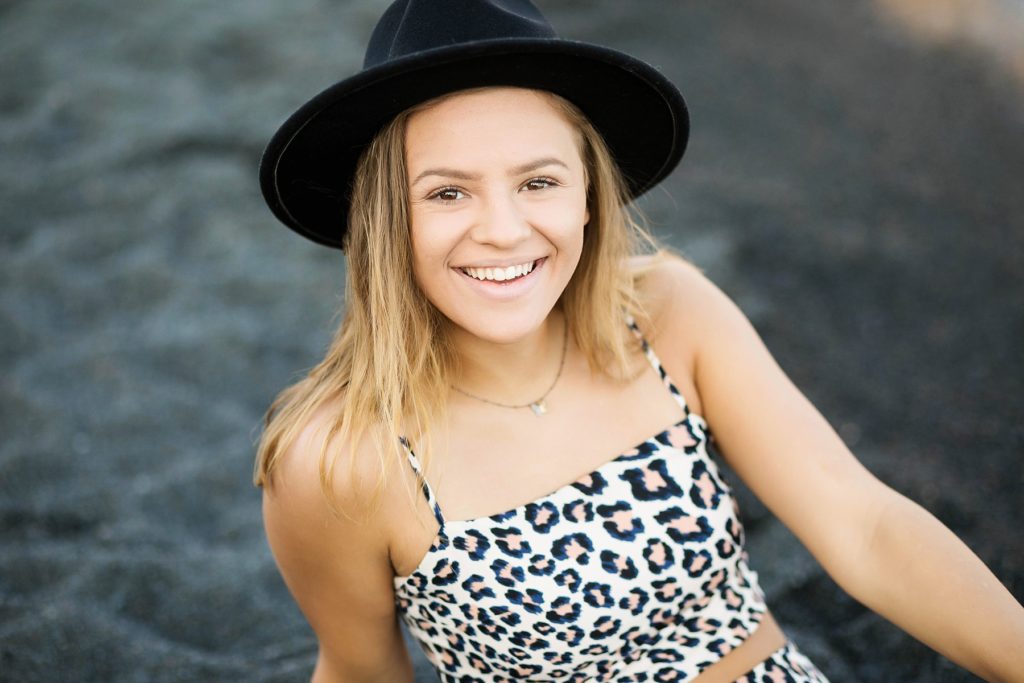girl sits in on a black sand beach on Lake Superior with a black hat on for her North Shore senior pictures