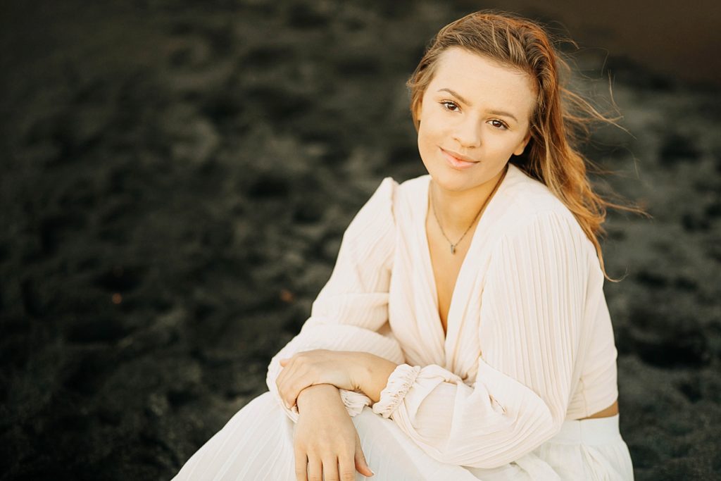girl sits in black sand on Black Sand Beach in Silver Bay. MN for her senior photos in a gown