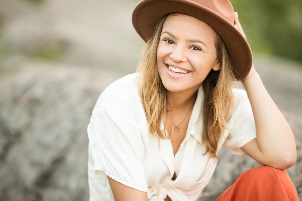 girl sits on a rock at Palisade Head for her senior photos on Lake Superior smiling with a brown hat on