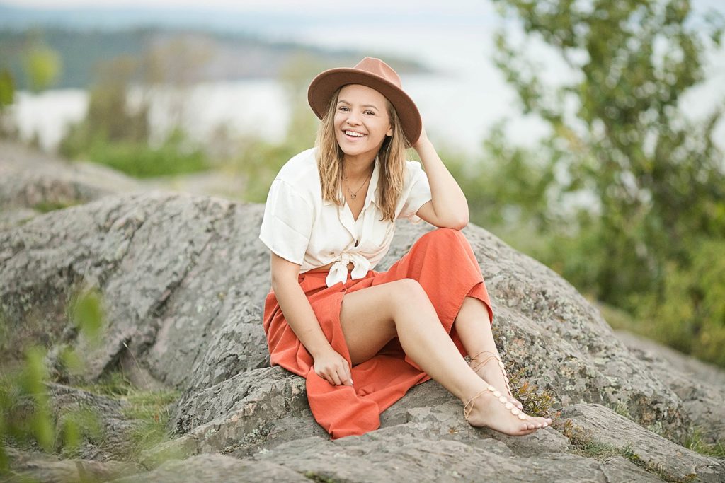 girl sits on a rock at Palisade Head for her senior photos on Lake Superior smiling with a brown hat on and a orange skirt