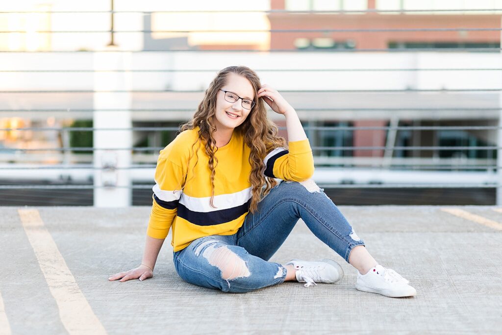 girl sitting on parking garage deck in yellow sweater smiling for her eau claire senior photos